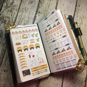 May include: A close-up of a black leather planner with colourful stickers. The stickers feature school-themed designs, including apples, buses, and a first day of school sign. There are also stickers with dental-themed designs, including teeth, toothbrushes, and a dentist.