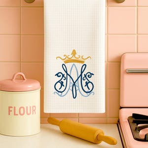 May include: White waffle-weave kitchen towel with a navy blue and light blue monogram design topped with a gold crown. A wooden rolling pin and a flour canister are in the foreground.