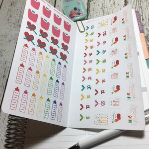 Op de afbeelding: Een set kleurrijke stickers voor een planner of dagboek. De stickers bevatten waterflesiconen, harten met meetlint en pijlen met nummers. De stickers zijn ontworpen om mensen te helpen hun fitnessdoelen bij te houden.