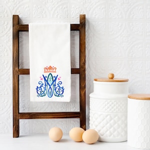 May include: White kitchen towel with a decorative design featuring a crown and the letter "M" in blue, green, and orange. The towel is hanging on a wooden ladder-style rack. Two white ceramic jars with wooden lids are visible.