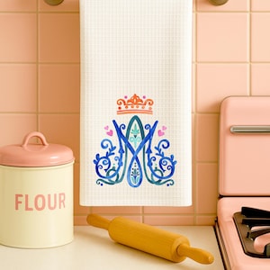 May include: White waffle-weave kitchen towel with a blue and teal monogram and crown design. The towel hangs on a gold-coloured bar. A pink flour canister and rolling pin are also visible in the kitchen setting.