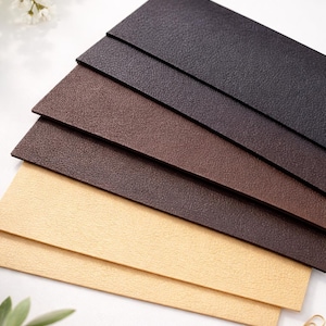 May include: A stack of textured paper in shades of brown and tan. The colors range from dark brown to light tan, with a slight grain visible on the surface. A gold paperclip rests on the white surface.