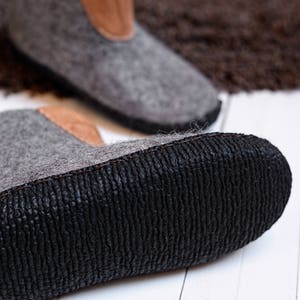 Puede incluir: Primer plano de dos botas de fieltro gris con detalles de cuero marr&oacute;n en la parte superior. Las botas tienen suelas de goma negra con un patr&oacute;n texturizado. El fondo est&aacute; borroso, con una alfombra marr&oacute;n y una superficie de madera blanca.
