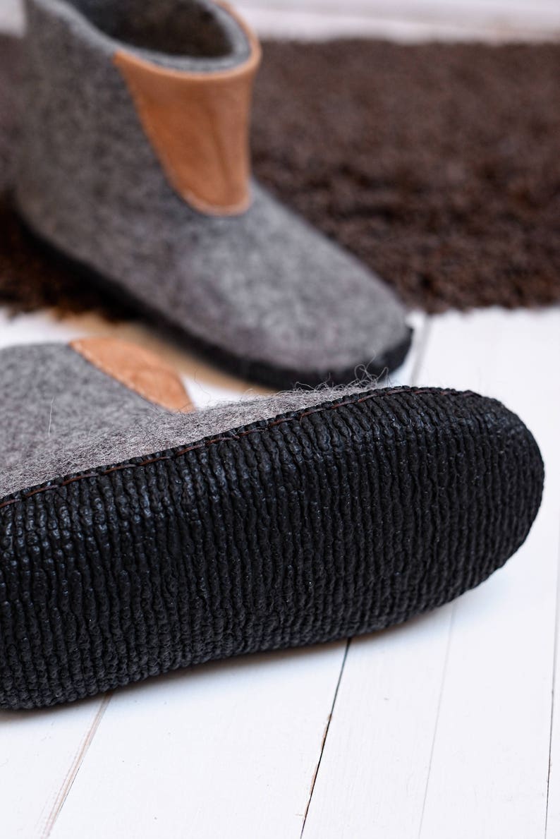 Puede incluir: Primer plano de dos botas de fieltro gris con detalles de cuero marr&oacute;n en la parte superior. Las botas tienen suelas de goma negra con un patr&oacute;n texturizado. El fondo est&aacute; borroso, con una alfombra marr&oacute;n y una superficie de madera blanca.