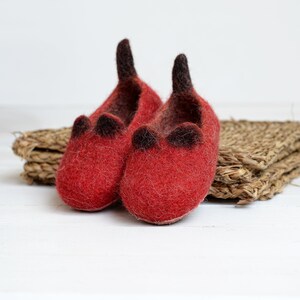 May include: A pair of red wool slippers with black felt accents. The slippers are sitting on a woven straw mat.