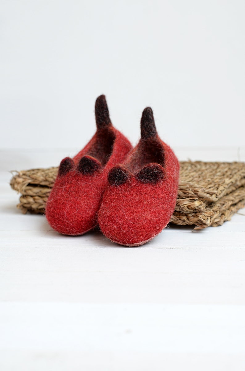 May include: A pair of red wool slippers with black felt accents. The slippers are sitting on a woven straw mat.