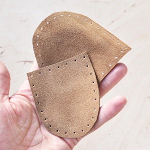May include: Two brown leather pieces with pre-punched holes, held in a hand. The pieces are in a teardrop shape, likely for crafting or sewing projects. The leather has a textured, suede-like appearance.