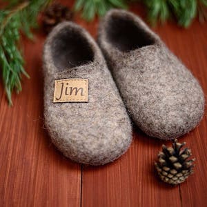 May include: A pair of grey wool slippers with a brown leather label that says "Jim". The slippers are on a wooden surface with pine needles and a pine cone.