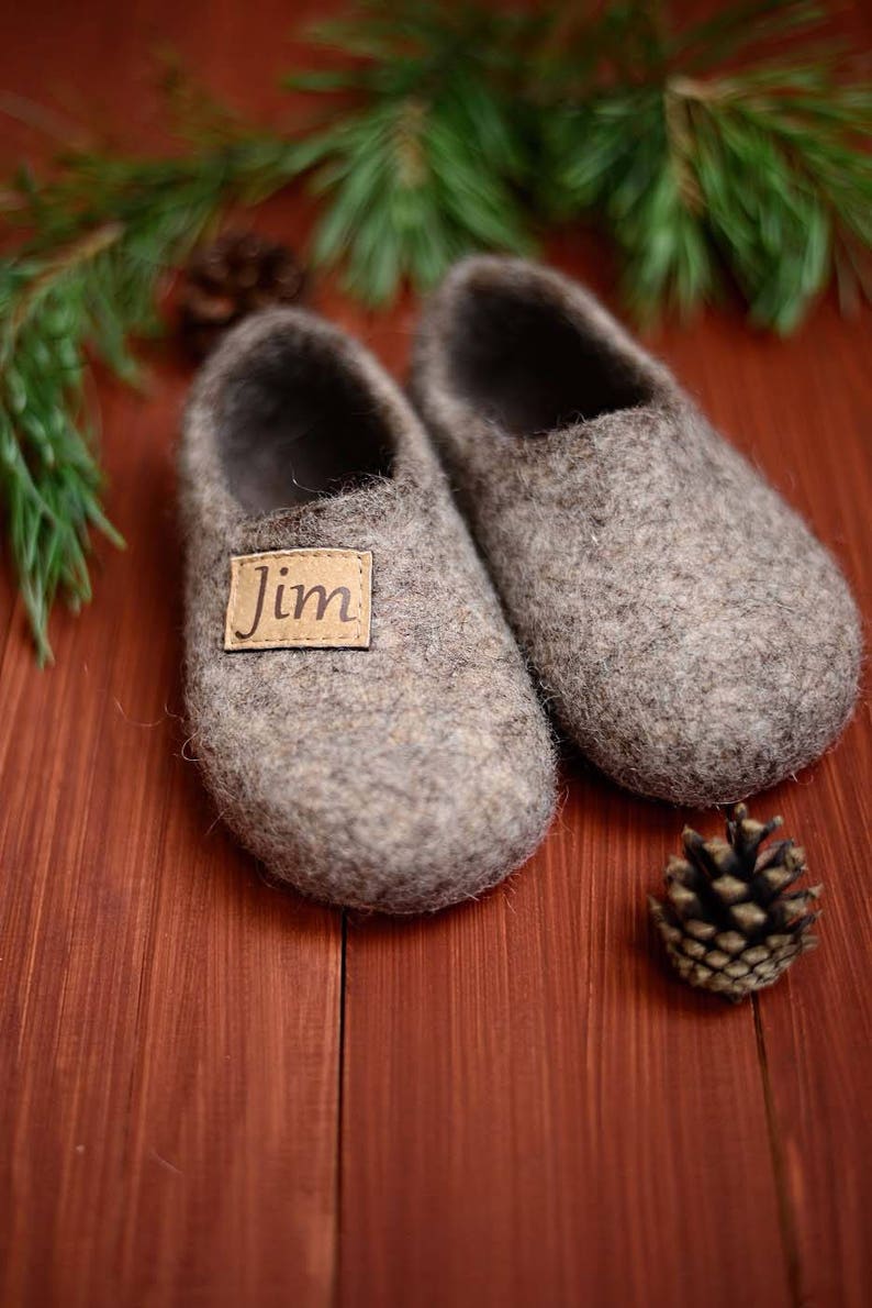 May include: A pair of grey wool slippers with a brown leather label that says "Jim". The slippers are on a wooden surface with pine needles and a pine cone.