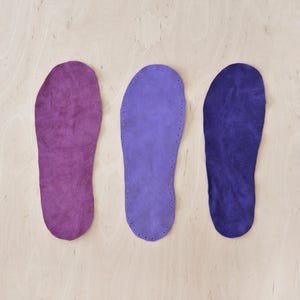 Op de afbeelding: Drie suède binnenzolen in tinten paars, lavendel en blauw liggen op een licht houten oppervlak. De middelste binnenzool heeft een rij gelijkmatig verdeelde gaten langs de rand, wat wijst op een stikpatroon. De binnenzolen zijn ontworpen voor de voetvorm.
