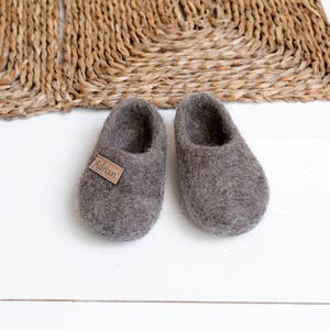 May include: A pair of grey wool slippers with a brown leather tag that says "Adrian". The slippers are on a white wooden surface.