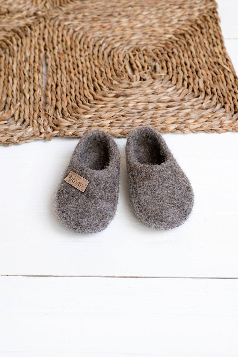 May include: A pair of grey wool slippers with a brown leather tag that says "Adrian". The slippers are on a white wooden surface.