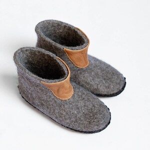 Puede incluir: Un par de zapatillas de fieltro gris con un adorno de cuero marr&oacute;n en el tobillo. Las zapatillas tienen una punta redondeada y una suela negra. Estas acogedoras zapatillas son perfectas para uso en interiores.
