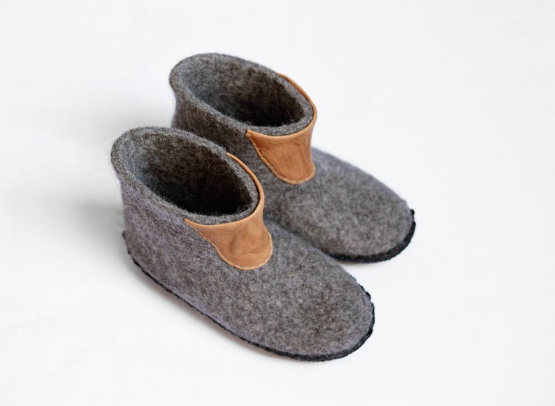 Puede incluir: Un par de zapatillas de fieltro gris con un adorno de cuero marr&oacute;n en el tobillo. Las zapatillas tienen una punta redondeada y una suela negra. Estas acogedoras zapatillas son perfectas para uso en interiores.