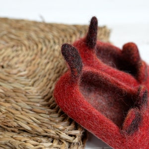 May include: Red felt slippers with black felt horns. The slippers are sitting on a woven straw mat.
