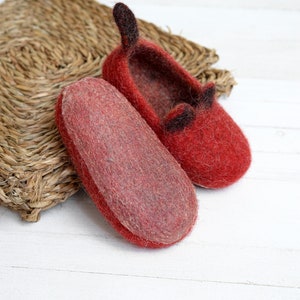 May include: A pair of red felt slippers with black felt ears. The slippers are on a white wooden surface.