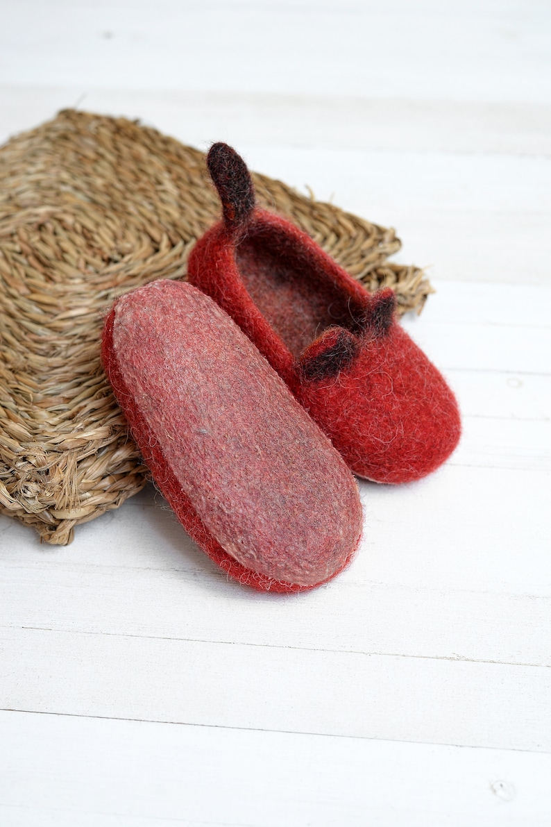 May include: A pair of red felt slippers with black felt ears. The slippers are on a white wooden surface.