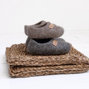 May include: Two pairs of grey wool slippers with leather labels. The slippers are stacked on top of each other and are sitting on a woven straw mat.