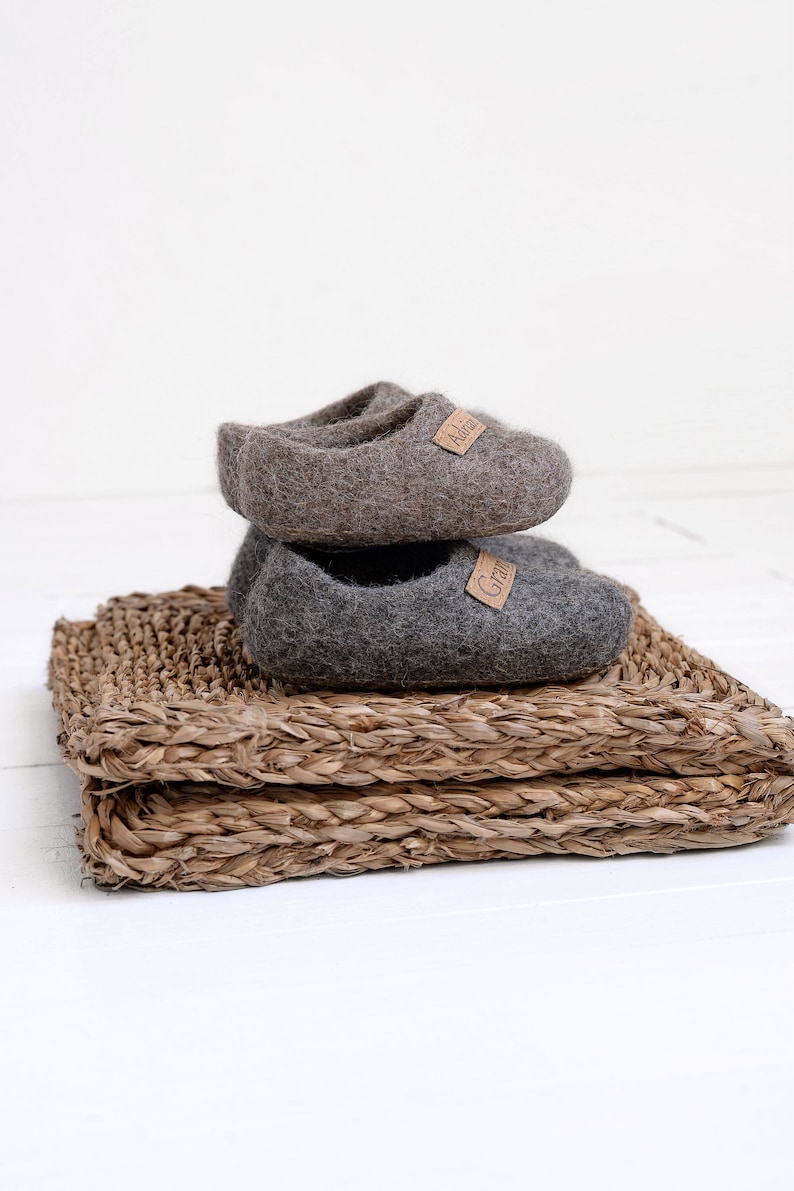 May include: Two pairs of grey wool slippers with leather labels. The slippers are stacked on top of each other and are sitting on a woven straw mat.