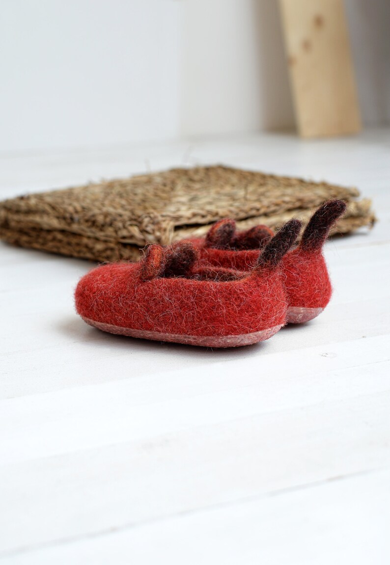 May include: A pair of red felt slippers with brown felt accents. The slippers have a pointed toe and a small heel.