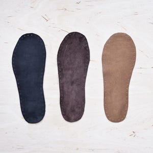 May include: Three leather insoles in navy, dark brown, and tan, displayed side by side. Each insole is foot-shaped, with small holes around the edges. The insoles are on a light-coloured wooden surface.