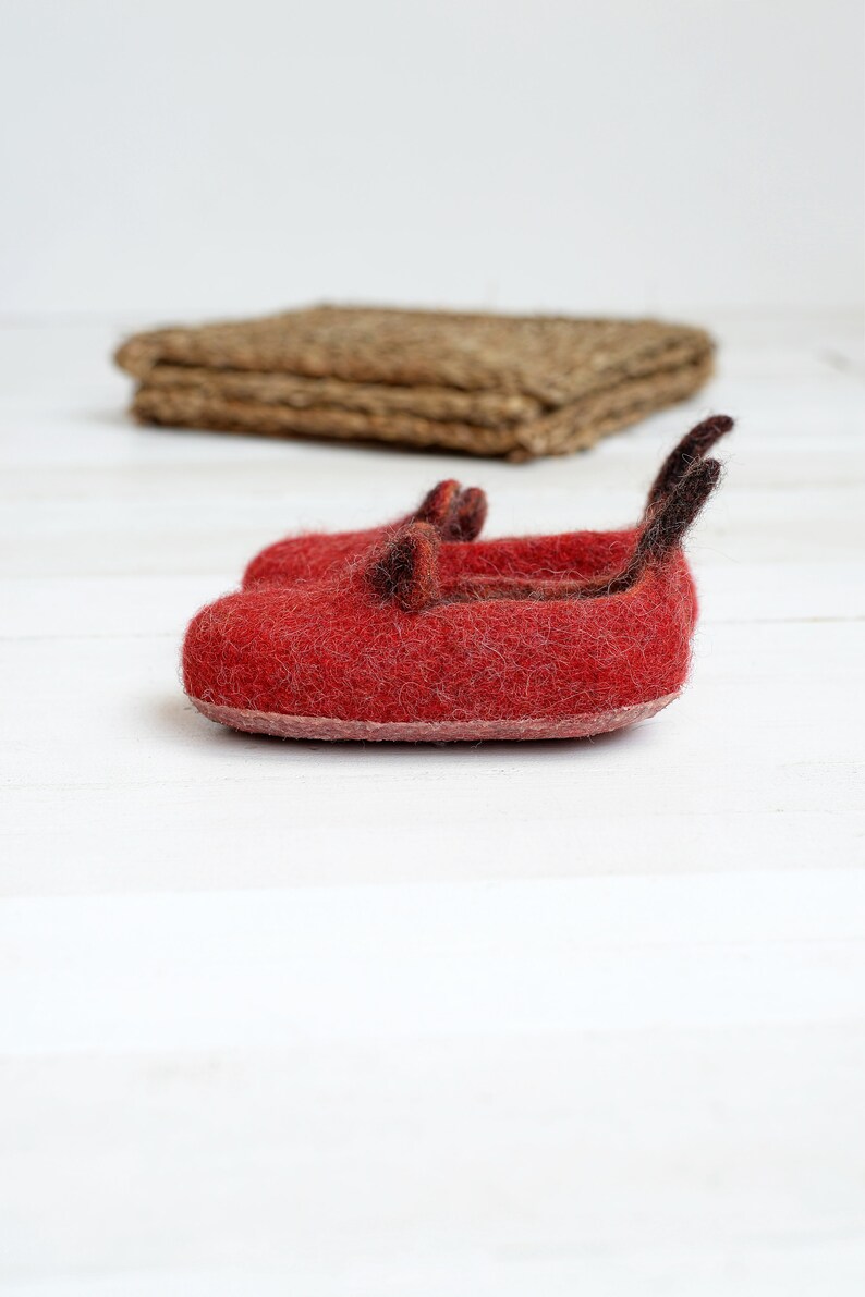 May include: A pair of red felt slippers with black felt accents. The slippers are on a white surface.