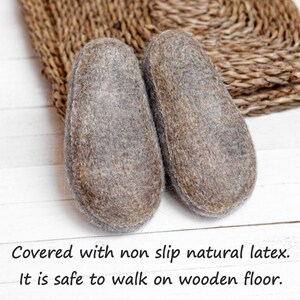 May include: A pair of grey felt slippers with a non-slip natural latex sole. The slippers are on a white wooden surface with a woven mat behind them. "Covered with non slip natural latex. It is safe to walk on wooden floor." is written below the slippers.