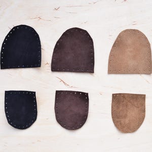 May include: Six leather pieces in various shades of brown and black, arranged on a light wood surface. Each piece has a rounded top and is pre-punched with holes for stitching. The leather pieces are in different shapes and sizes.