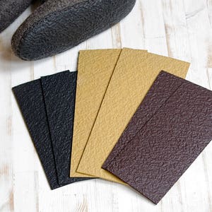 Op de afbeelding: Een paar grijze vilten pantoffels met zwarte rubberen zolen bevinden zich boven een waaier van getextureerde rechthoekige inlegzolen in zwart, beige en bruin. De inlegzolen zijn ontworpen voor schoeisel.