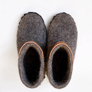 Puede incluir: Un par de zapatillas de fieltro gris con un acento de cuero sobre un fondo blanco. Las zapatillas tienen una punta redondeada y un ribete cosido negro. El interior de las zapatillas es gris oscuro.