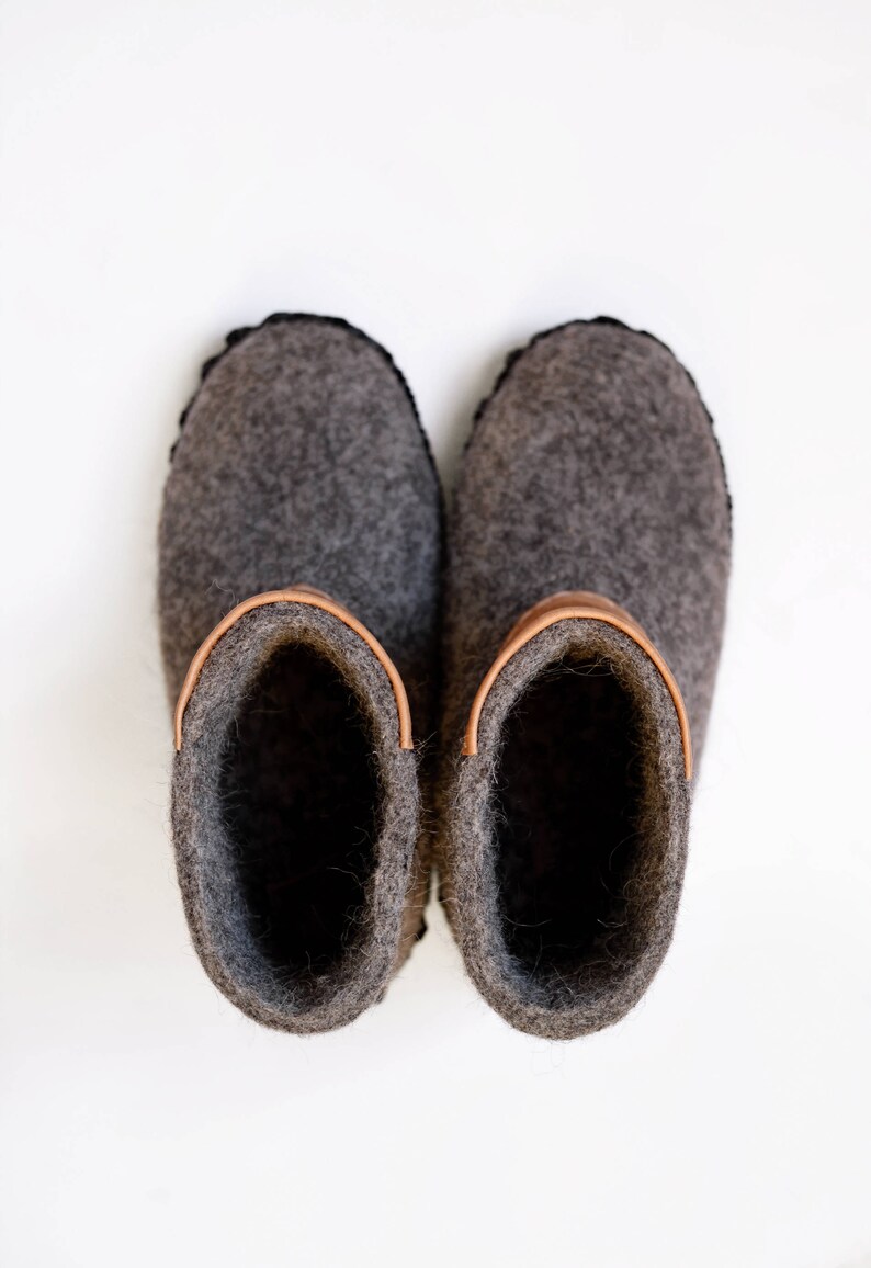 Puede incluir: Un par de zapatillas de fieltro gris con un acento de cuero sobre un fondo blanco. Las zapatillas tienen una punta redondeada y un ribete cosido negro. El interior de las zapatillas es gris oscuro.