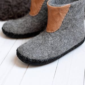 Puede incluir: Un par de botas de lana afieltrada gris con un acento de cuero marr&oacute;n en la parte superior. Las botas tienen una suela cosida negra y est&aacute;n dise&ntilde;adas para uso en interiores. Las botas se exhiben sobre una superficie de madera blanca.