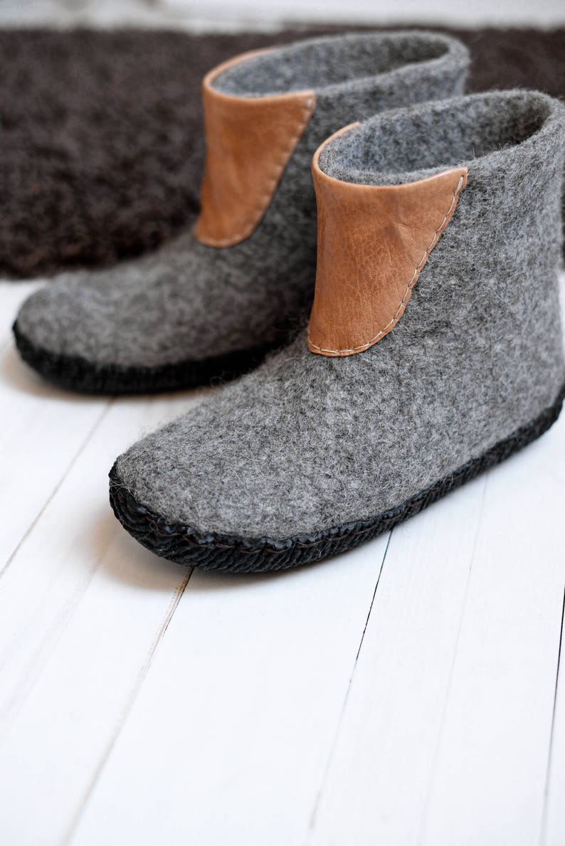 Puede incluir: Un par de botas de lana afieltrada gris con un acento de cuero marr&oacute;n en la parte superior. Las botas tienen una suela cosida negra y est&aacute;n dise&ntilde;adas para uso en interiores. Las botas se exhiben sobre una superficie de madera blanca.
