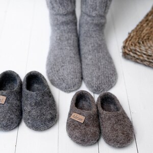 May include: Two pairs of grey wool slippers with personalised labels. The slippers are on a white wooden floor. One pair has the name "Adrian" on the label and the other pair has the name "Ant" on the label.
