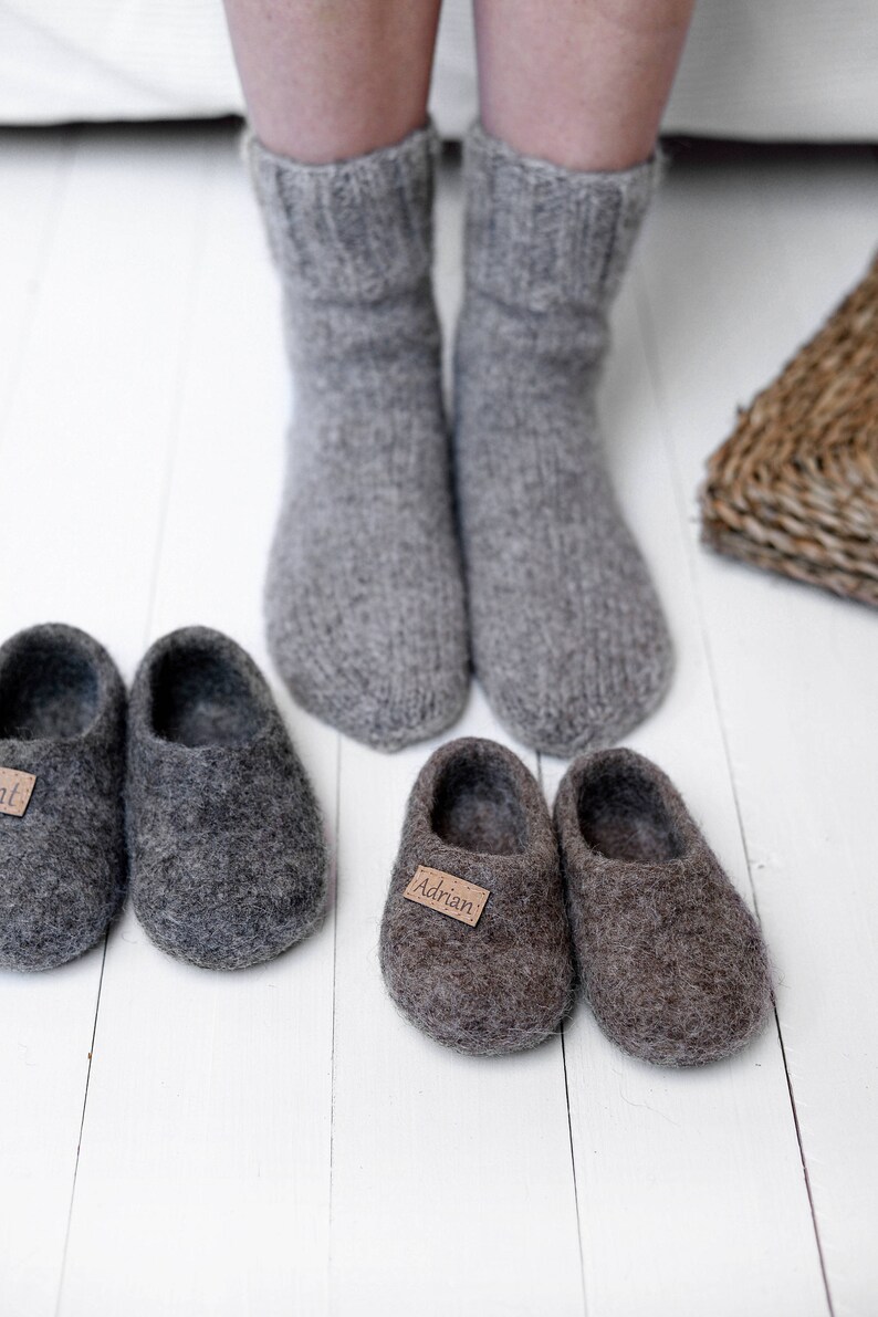 May include: Two pairs of grey wool slippers with personalised labels. The slippers are on a white wooden floor. One pair has the name "Adrian" on the label and the other pair has the name "Ant" on the label.