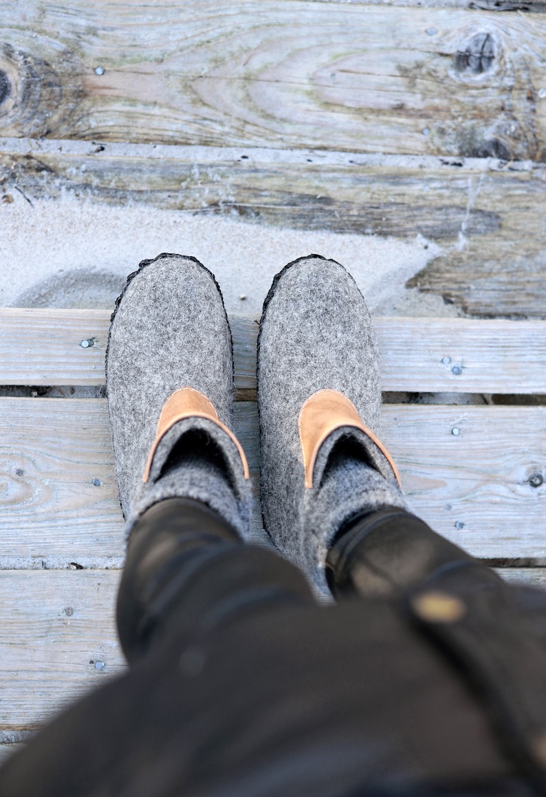 Puede incluir: Un par de zapatillas grises de fieltro con un detalle de cuero marr&oacute;n claro en la parte superior. Las zapatillas tienen un ribete negro y se ven desde arriba, con las piernas del usuario visibles.