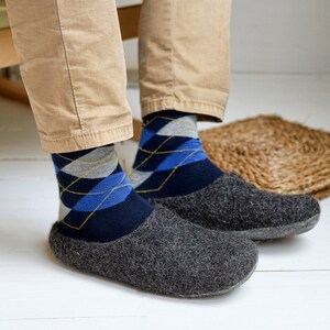 May include: A person wearing argyle socks and gray felt slippers. The socks are navy blue with blue, gray, and gold diamond patterns. The slippers are a dark gray, wool-like material.