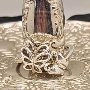 May include: A silver-toned ring featuring two butterfly motifs. One butterfly is designed with an open-work, filigree-style outline, while the adjacent butterfly is solid and smooth. The ring is displayed on a reflective, ornate silver-toned stand.