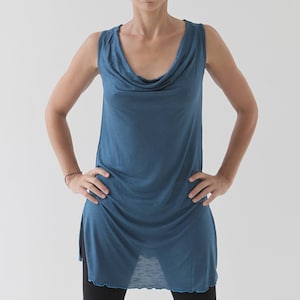 May include: A woman wearing a teal blue sleeveless tunic top with a cowl neck. The top has a slightly sheer fabric and a scalloped hem.