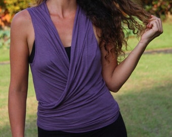 May include: A sleeveless purple top featuring a deep V-neckline with a draped, wrap-style front design. The garment is made of a soft, lightweight fabric and is worn over a black base layer, paired with solid black pants for a simple look.
