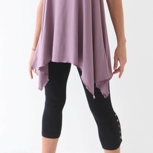 May include: A woman wearing a loose-fitting, asymmetrical, mauve-colored top with a black pair of leggings that have decorative buttons on the side.