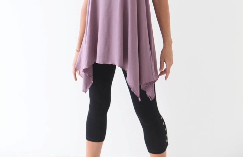 May include: A woman wearing a loose-fitting, asymmetrical, mauve-colored top with a black pair of leggings that have decorative buttons on the side.