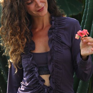 May include: A woman wearing a long-sleeved, purple, ruffled blouse with a tie at the waist. She is holding a small pink flower in her hand.