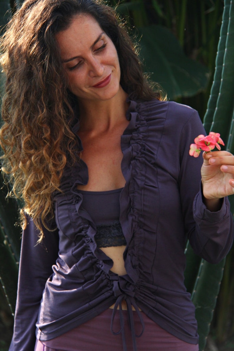 May include: A woman wearing a long-sleeved, purple, ruffled blouse with a tie at the waist. She is holding a small pink flower in her hand.