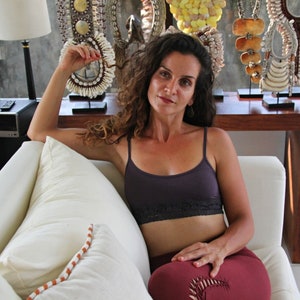 May include: A woman wearing a purple tank top with black lace trim sits on a white couch. She is wearing maroon pants and has curly brown hair. She is holding a white shell necklace in her hand. The couch is in front of a window with a wooden table and several shell necklaces on display.