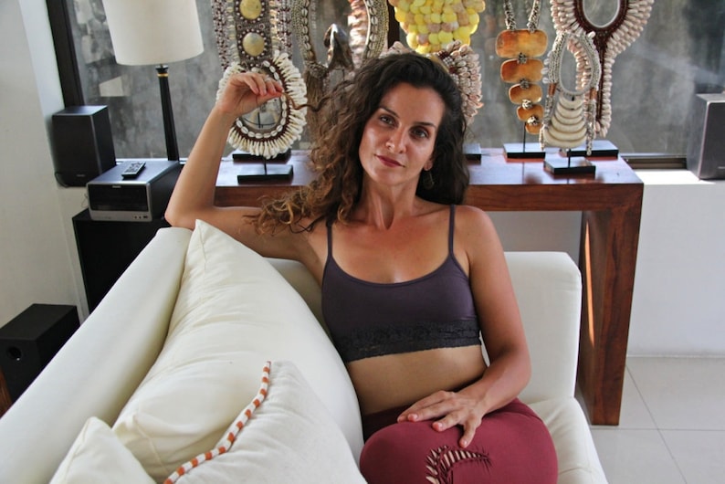 May include: A woman wearing a purple tank top with black lace trim sits on a white couch. She is wearing maroon pants and has curly brown hair. She is holding a white shell necklace in her hand. The couch is in front of a window with a wooden table and several shell necklaces on display.
