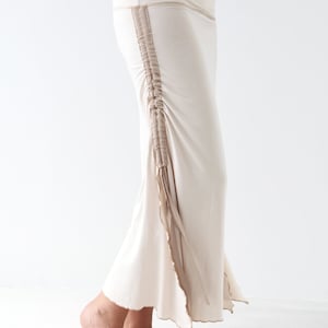 May include: A long, flowing skirt in a light beige color with a brown trim along the side. The skirt has a gathered detail at the side and a flowing, asymmetrical hem.