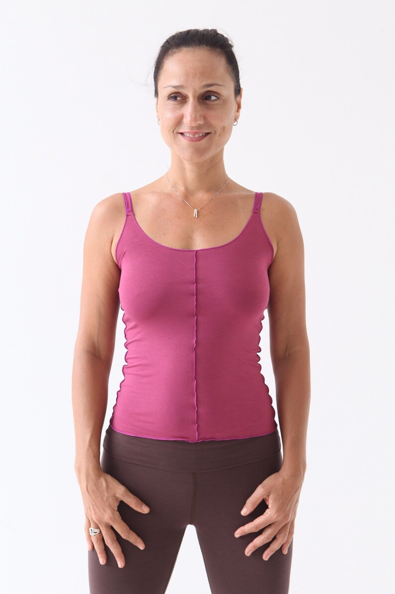 May include: A woman wearing a pink tank top and brown leggings. She is standing with her hands on her hips and looking to the side.