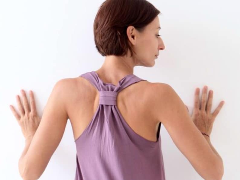 May include: A woman wearing a purple tank top with a knotted back detail, standing with her hands on a white wall.