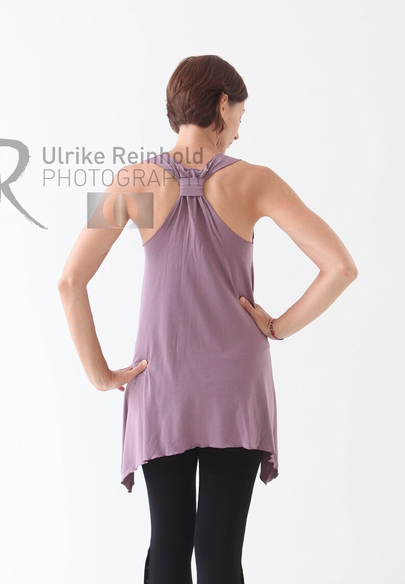 May include: A woman wearing a mauve tank top with a knotted back detail and black leggings. The top has a slightly asymmetrical hemline.